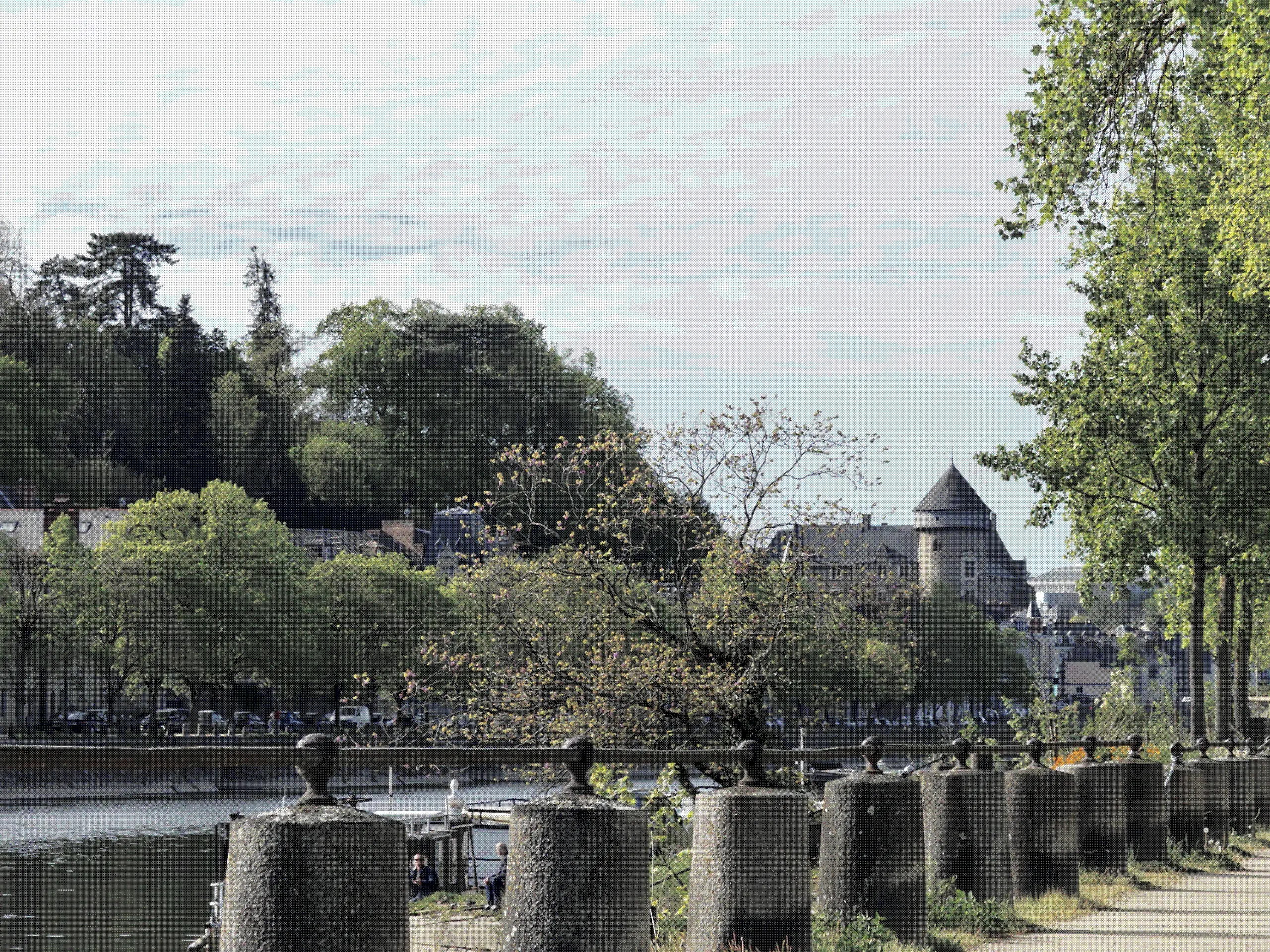 Picture of a riverside path, with a medieval castle in the background and trees all around. The sky is covered by clouds amd the sun is shining.